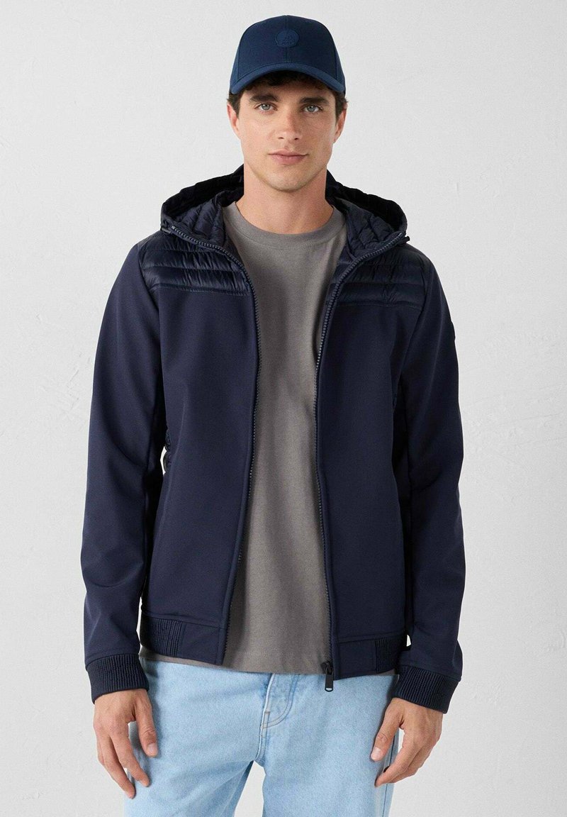 Marineblauwe gewatteerde hoodie met gestructureerd bovenpaneel, ritsafsluiting, geribde manchetten, gecombineerd met een grijs shirt en lichtblauwe jeans.