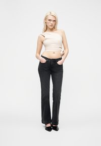 Νεαρή γυναίκα με μακριά ξανθά μαλλιά, που φοράει μπεζ τοπ ενός ώμου, μαύρο τζιν και μαύρα μυτερά παπούτσια, με τα χέρια στις τσέπες.