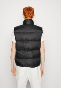 Svart vadderad väst med hög krage och ärmlös design. Slät tygtextur med synlig horisontell sömnad. Enkelt bakifrån.