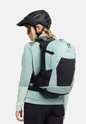 Uno zaino verde menta e nero con spallacci regolabili, dettagli in rete e tasche con cerniera, indossato sopra una maglietta azzurro chiaro e pantaloni corti.