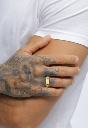 Tätowierte Hand mit einem goldenen Ring, die sanft auf dem Oberarm einer Person mit weißem Kurzarmhemd ruht.