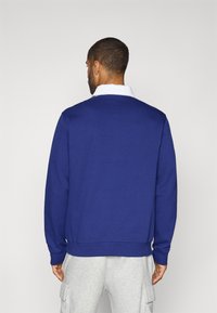 Marineblå sweater med lange ærmer, ribbede manchetter og kant, med en hvid krave skjorte synlig ved halsen. Ensartet tekstur, afslappet pasform.