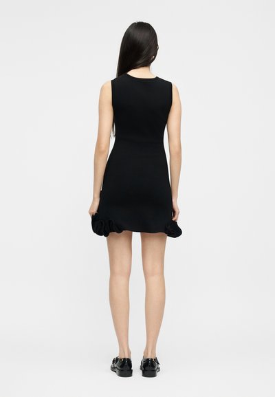 Γυναίκα που στέκεται με την πλάτη στην κάμερα, φοράει μαύρο μίνι φόρεμα χωρίς μανίκια με βολάν στο τελείωμα και μαύρα μοκασίνια, σε απλό γκρι φόντο.