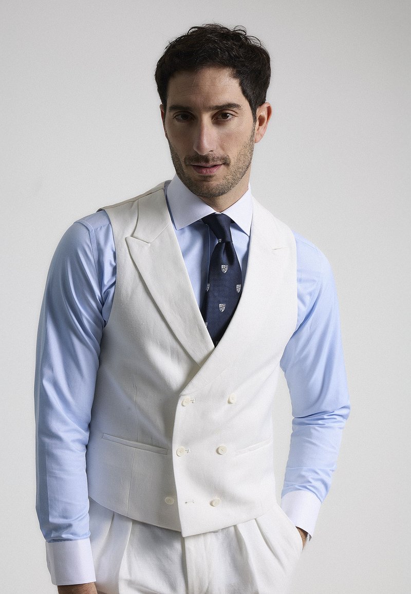 Chaleco blanco de doble botonadura con una textura suave, llevado sobre una camisa celeste y una corbata oscura con un patrón. Diseño sencillo y a medida.