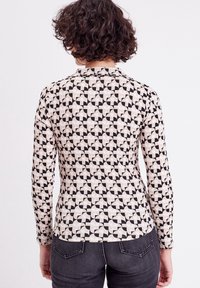 Mujer con cabello corto y rizado, vistiendo una blusa de manga larga con un patrón geométrico y jeans oscuros, de espaldas frente a un fondo blanco liso.