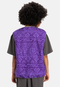 Fialová vzorovaná vesta s geometrickými dizajnmi, uvoľneného strihu a krátkymi rukávmi, nosená cez šedú košeľu. Textúrovaný povrch látky.