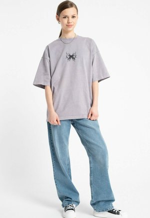 Młoda kobieta w oversize'owej szarej koszulce z czarnym wzorem, w jasnoniebieskich szerokich dżinsach, w czarno-białych sneakersach, stojąca na tle białego tła.