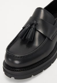 Mocasines de cuero negro con un acento de borlas, punta redondeada y suela de goma gruesa. Textura suave y costuras detalladas a lo largo de los bordes.