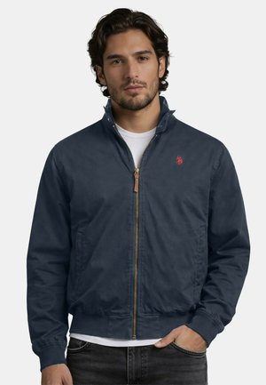 Mann mit dunklem welligem Haar, der eine marineblaue Jacke mit Reißverschluss über einem weißen Hemd trägt, steht vor einem einfarbigen hellen Hintergrund.