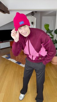 Red knit beanie with a branded patch, burgundy and pink geometric-patterned zip-up sweater, black trousers, and silver sneakers.