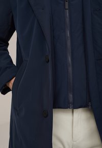 Chaqueta aislante azul marino con una textura suave, con cremallera frontal y dos botones, superpuesta a una camisa más clara.