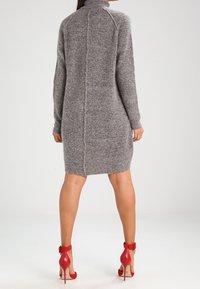 Abito maglione grigio con alto colletto, texture morbida, polsini a costine e dettagli di cucitura centrale. Abbinato a scarpe con tacco rosso.