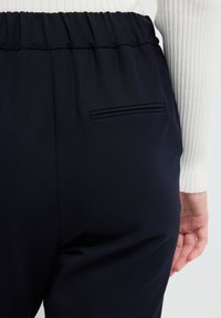 Navyblauwe broek met een elastische tailleband, voorzien van een achterzakdetail en een gladde, textuurachtige stofafwerking.