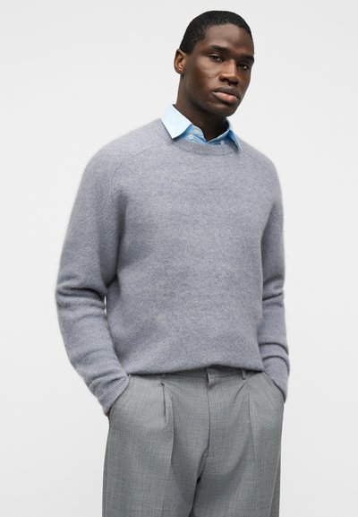Άνδρας που φοράει ανοιχτόγαλάζιο πουκάμισο με γιακά κάτω από γκρι χνουδωτό πουλόβερ και γκρι πιετέ παντελόνια, με τα χέρια στις τσέπες, μπροστά σε λευκό φόντο.