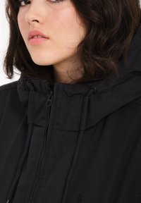 Schwarze Jacke mit hohem Kragen, ausgestattet mit einem Reißverschluss und Kordeln. Der Stoff wirkt strukturiert und strapazierfähig, mit dezenten Details.