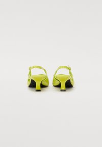 Escarpins en cuir verni jaune avec une petite base noire, partie arrière fermée, bride ajustable à la cheville et un design minimaliste.