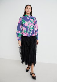 Blomstrete bluse i lilla, rosa og grønn; har knapper foran og lange puffærmer. Svart, lagdelt tyllskjørt med glitter. Svart flate sko.