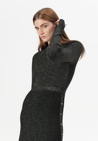 Modella in un abito nero glitterato che assume una posa sicura, con una mano che tocca il collo e un'espressione seria.