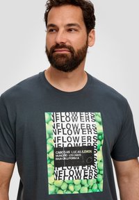 Grijze t-shirt met korte mouwen, voorzien van een grote grafische afbeelding met groene bloemenpatronen en zwarte tekstoverlay, die een textuurvol, modern ontwerp benadrukt.