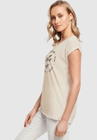 Beige bomulds T-shirt med korte ærmer, prydet med sort cirkulært tekstdesign på forsiden. Sammen med hvide bukser. Baggrunden er neutral grå.