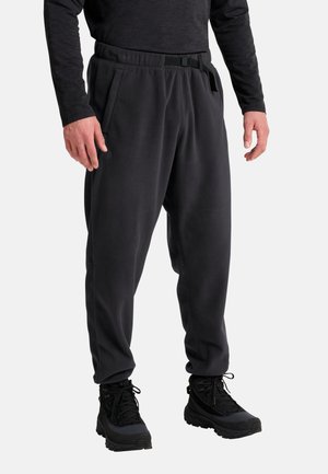Uomo che indossa una maglia nera a maniche lunghe, pantaloni in pile neri con tasche laterali e cintura incorporata, e scarponi da trekking neri, in piedi con le mani rilassate.