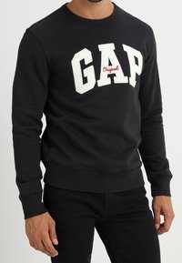 Sweat-shirt noir à encolure ronde, présentant un logo "GAP" blanc et une broderie rouge "Original", confectionné dans un tissu lisse.