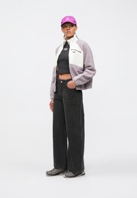 Fialovo-biela fleecová bunda, čierny krátky top, široké čierne kordurové nohavice a sivé tenisky s fialovou šiltovkou.