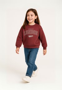 Felpa bordeaux con design testuale "LONDON" e "Welcome", abbinata a jeans blu e sneakers bianche. Tessuto morbido e vestibilità comoda.