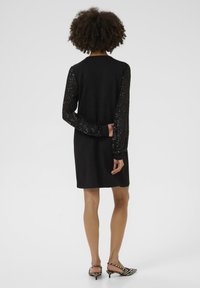 Crna haljina s dugim rukavima ukrašenim šljokicama, blago prilagođenog dizajna, do koljena, uparena s crno-bijelim prugastim sandalama na visoku petu.