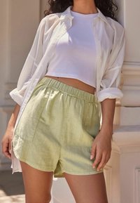 Chemise blanche boutonnée portée sur un débardeur blanc court et un short vert clair en mélange de lin, avec une taille élastique et des poches latérales.