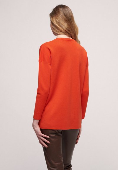 Oversized orange knit sweater with long sleeves, scoop neckline, and ribbed texture. Worn with brown fitted pants, showing a relaxed fit.