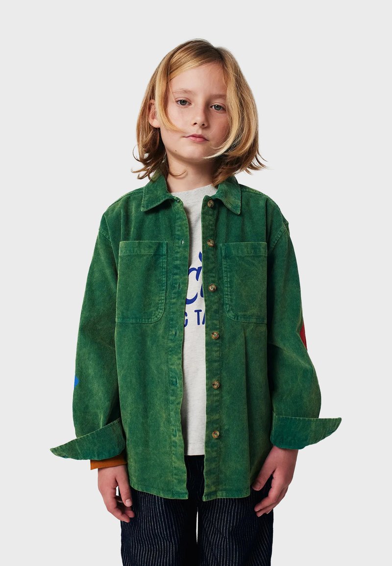 Camisa de pana verde con bolsillos de solapa, botones en la parte delantera y mangas de color contrastante. Llevada con una camiseta gris y pantalones a rayas.