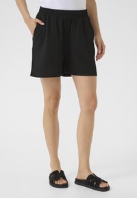 Shorts noirs avec une taille élastique, tissu texturé et poches latérales. Associés à des sandales noires à glissière avec un design de bride.