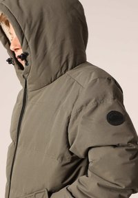 Veste matelassée verte avec capuchon, dotée d'une fermeture éclair, de poches latérales, d'un tissu texturé et d'un écusson rond noir sur la manche.