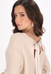 Maglione in maglia beige chiaro con un motivo testurizzato, schiena scoperta e un dettaglio a fiocco in tessuto. Il modello mostra un profilo laterale con onde morbide.