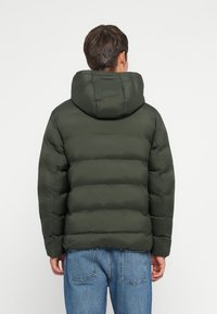 Uomo che indossa una giacca a vento verde scuro con cappuccio, di spalle, abbinata a jeans blu su uno sfondo bianco uniforme.