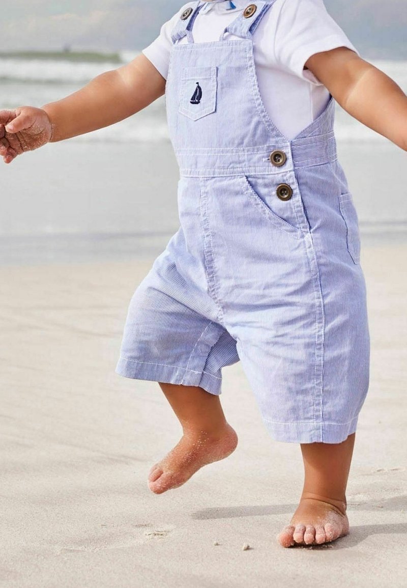 Tout-petit portant une salopette rayée bleu clair et une chemise blanche, marchant pieds nus sur une plage de sable près des vagues de l'océan.