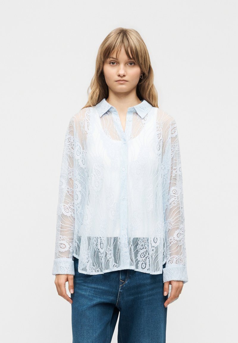 Young woman facing forward wearing a semi-transparent white lace button-up shirt over a collared base layer and blue jeans.