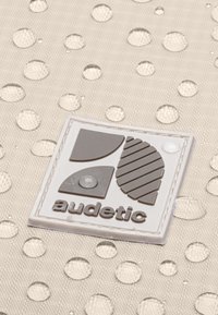 Logo quadrato bianco con forme geometriche grigie e testo "audetic", su uno sfondo beige texturizzato con motivi di gocce di pioggia.