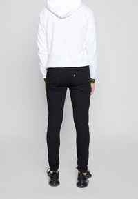 Svarta jeans med smal passform och en röd logotagg på bakfickan, bärs tillsammans med en vit hoodie och svarta sneakers.