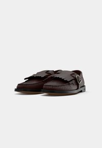 Chaussures en cuir brun avec une semelle plate, dotées d'un design frangé sur le dessus et d'une boucle métallique sur le côté.