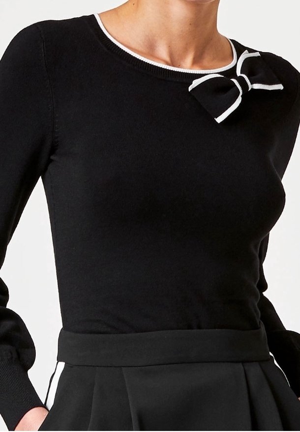 Pull noir en tissu tricoté, avec un col rond blanc et un petit nœud sur l'épaule gauche. Manches longues et ajustées.