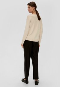 Beige Strickpullover mit langenÄrmeln, lockerer Passform und geripptem Saum, kombiniert mit schwarzer, weit geschnittenen Hose und schwarzen Schuhen. Einfarbiger Hintergrund.