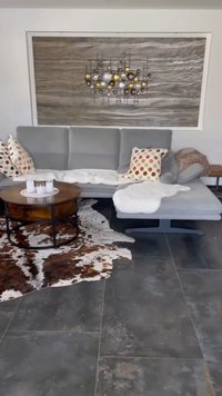 Canapea secțională gri cu perne cu buline și pături albe, masă de cafea rotundă din lemn cu lumânări, artă murală abstractă și covor din piele de vacă pe podeaua din gresie.