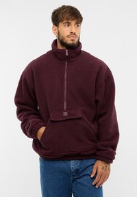 Burgundi fleece pulóver magas gallérral, félig cipzáros elülső résszel és kenguru zsebbel. Puha tapintású és laza fazonú. Logóval ellátva.