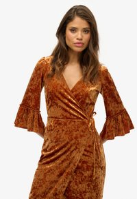 Robe portefeuille en velours couleur rouille avec des manches évasées, une finition texturée et un lien ajustable à la taille. Présente un décolleté en V profond.