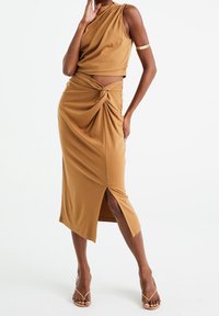 Hnědý zkrácený top s volným designem, který je spárován s midi sukní s uzlem na přední straně a bočním rozparkem, vyrobenou z jemného, pružného materiálu.