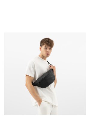 Młody mężczyzna w białym stroju trzymający czarną plecioną torbę na ramię na jednolitym jasnym tle.