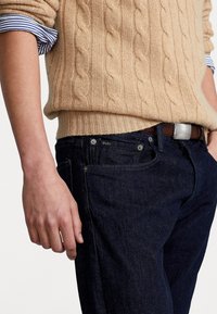Beige kabelstickad tröja över mörkblå jeans. Tröjan har en ribbad nederkant och ärmslut; jeansen har en sydd logotyp och silverdetaljer.