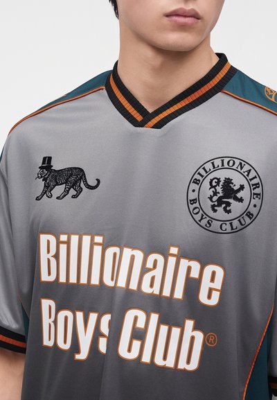 Νέος που φοράει γκρι και πράσινο μπλουζάκι Billionaire Boys Club με μαύρα λιοντάρι και λεοπάρδαλη λογότυπα και έντονο λευκό και πορτοκαλί κείμενο.